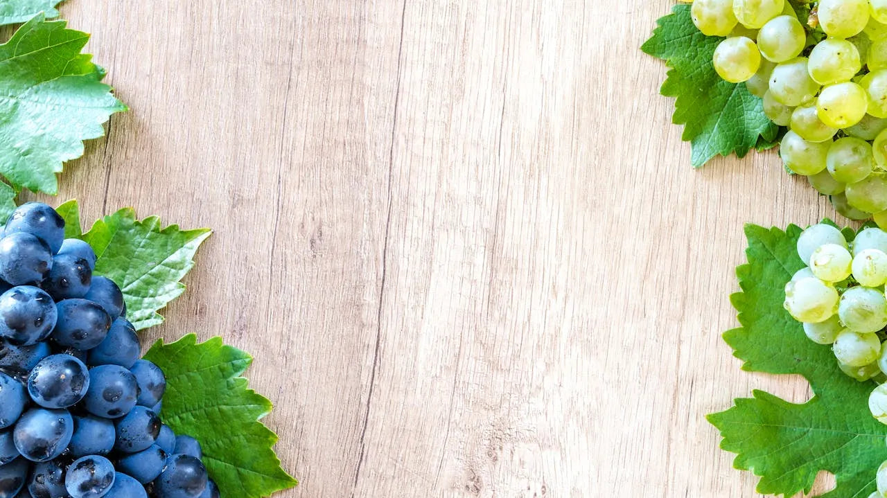 Et træbord dekoreret med blå og grønne drueklaser i hjørnerne, med grønne blade omkring druerne. Den centrale del af bordet er tom, hvilket giver en enkel og naturlig baggrund.