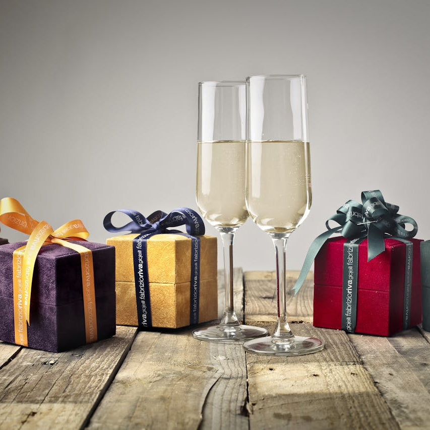 Fem farverige gaveæsker med bånd og to fyldte champagneglas stående på et rustikt træbord. Perfekt til festlige lejligheder og fejring