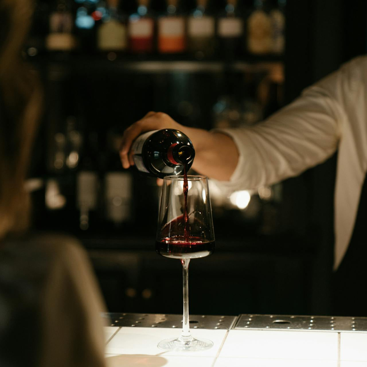 Person hælder rødvin i et glas på en oplyst bardisk med en baggrund af vinflasker og en dæmpet atmosfære.