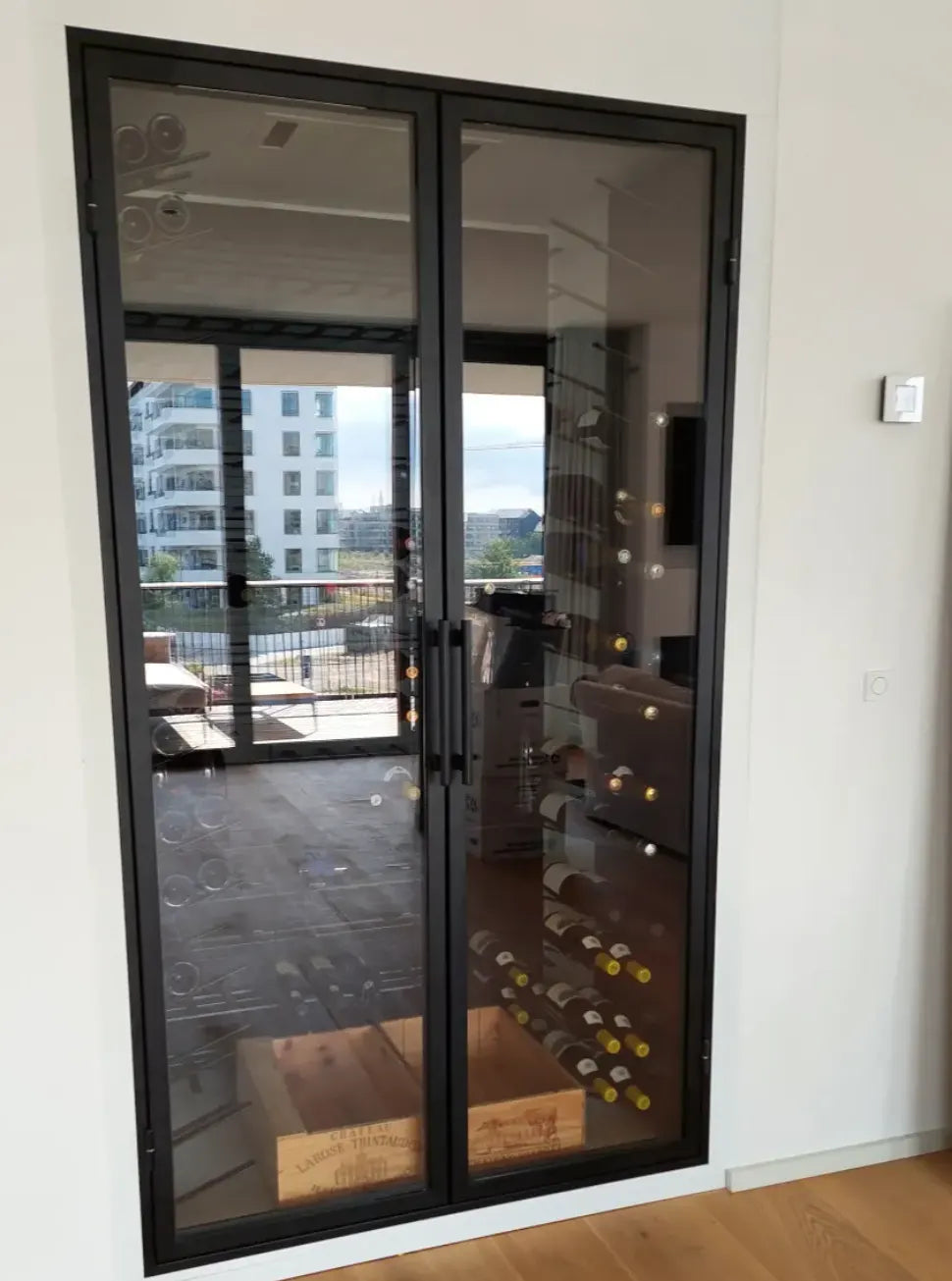Vinrum med glasdøre og sorte rammer, indeholdende flere vinflasker på vægmonterede holdere. Rummet har trækasser på gulvet og udsigt til en balkon og moderne bygninger i baggrunden.