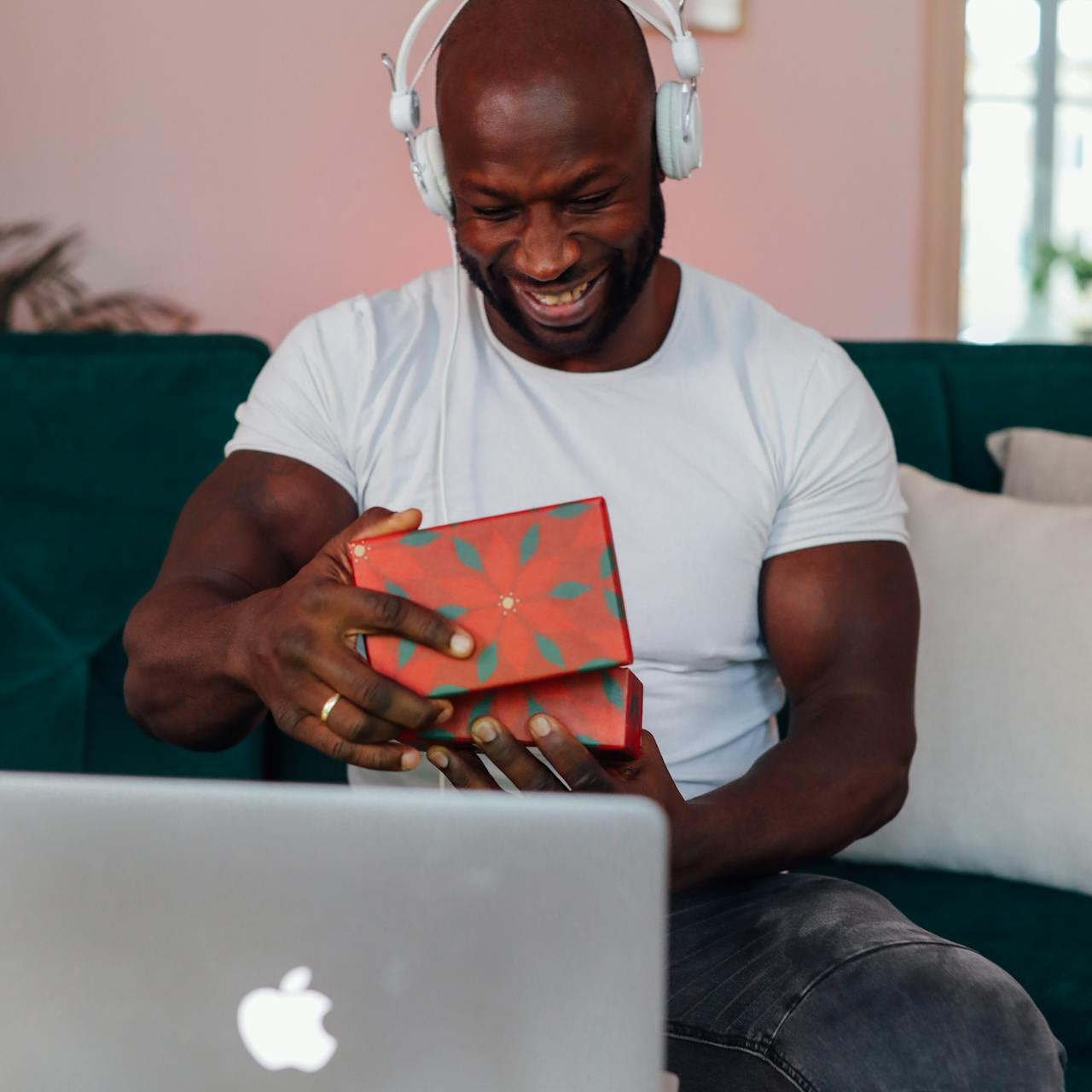 Mand iført høretelefoner og hvid t-shirt, der smilende åbner en gaveæske foran en bærbar computer. Han sidder på en grøn sofa i et hyggeligt hjem