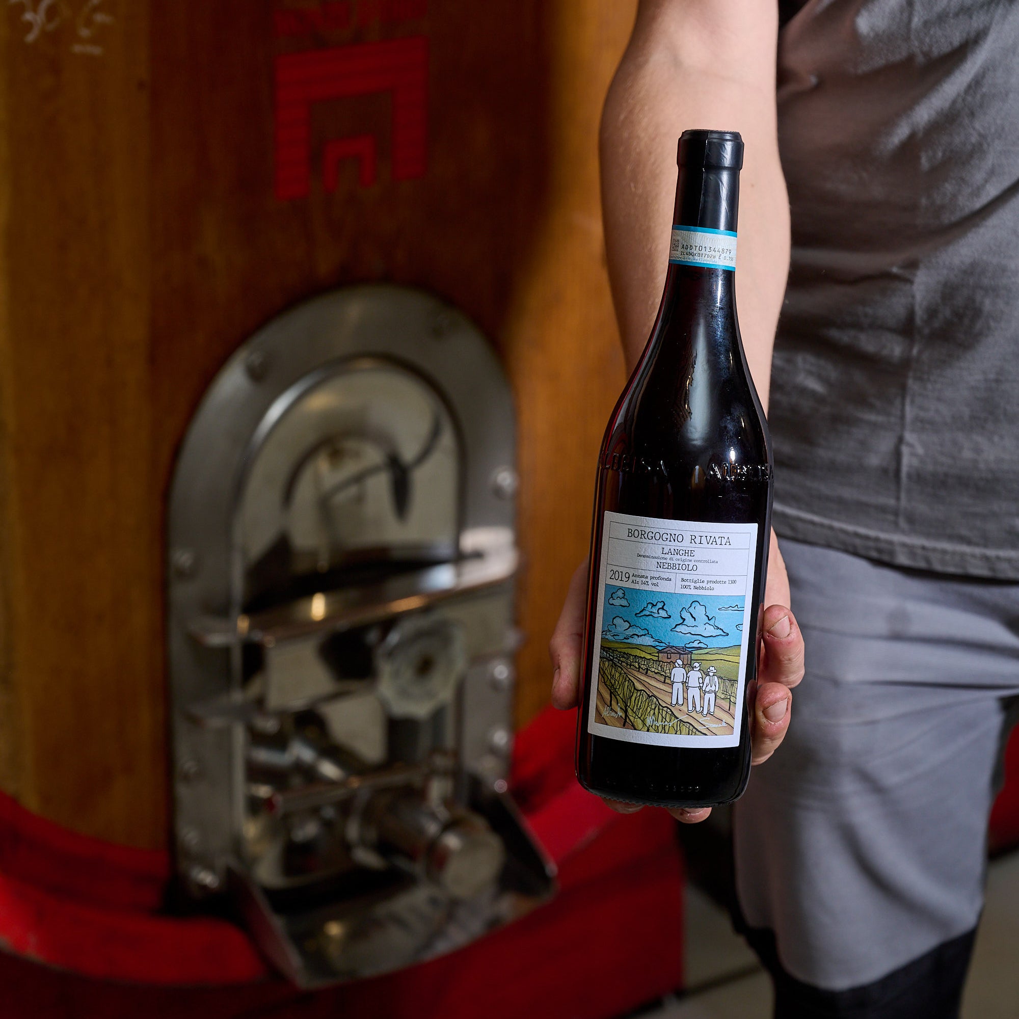 Person iført grå tøj holder en vinflaske foran en stor vintønde i en vinkælder. Fokus på flasken og etiketten.