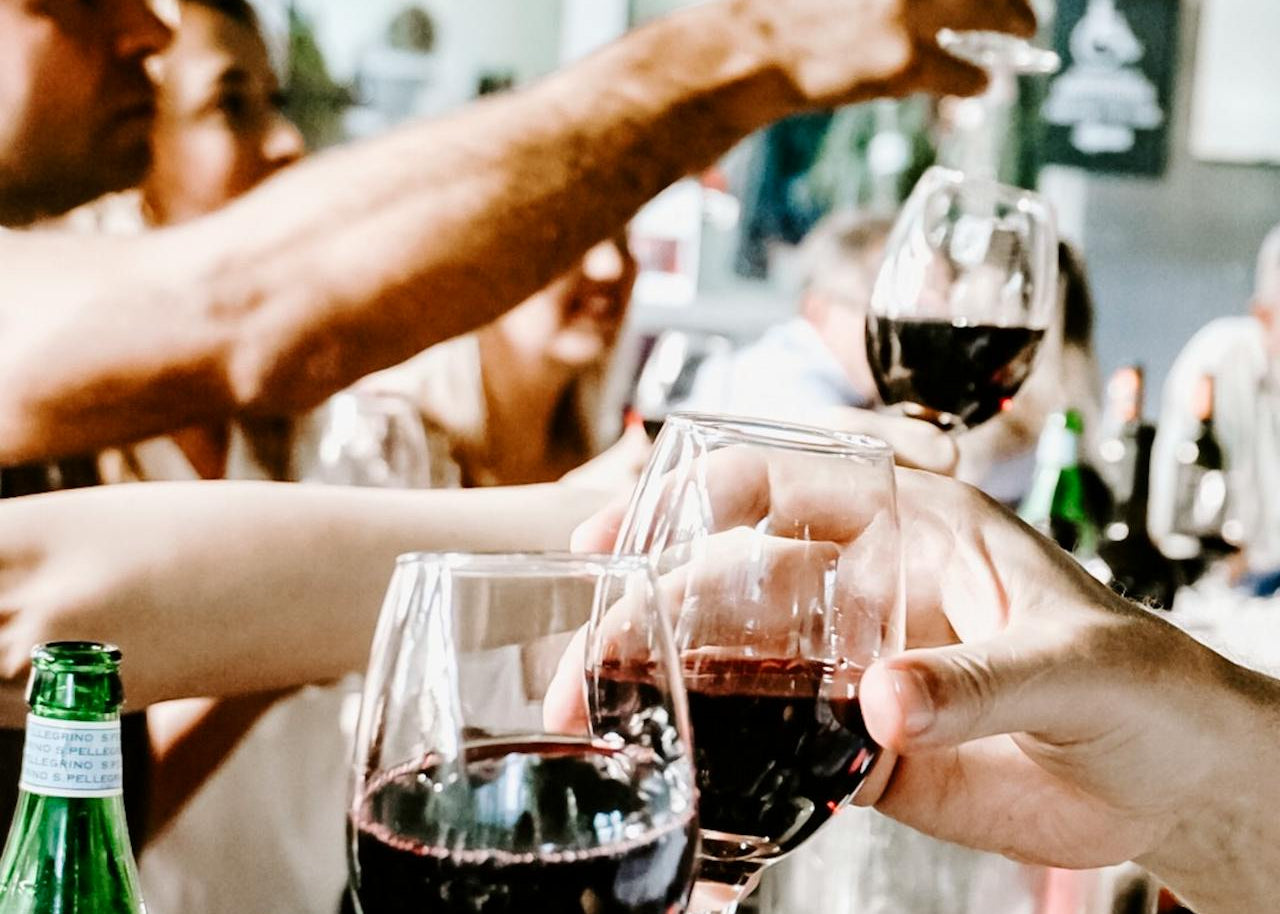 Gruppe mennesker skåler med glas rødvin rundt om et bord til en social sammenkomst, med flasker og mad i forgrunden