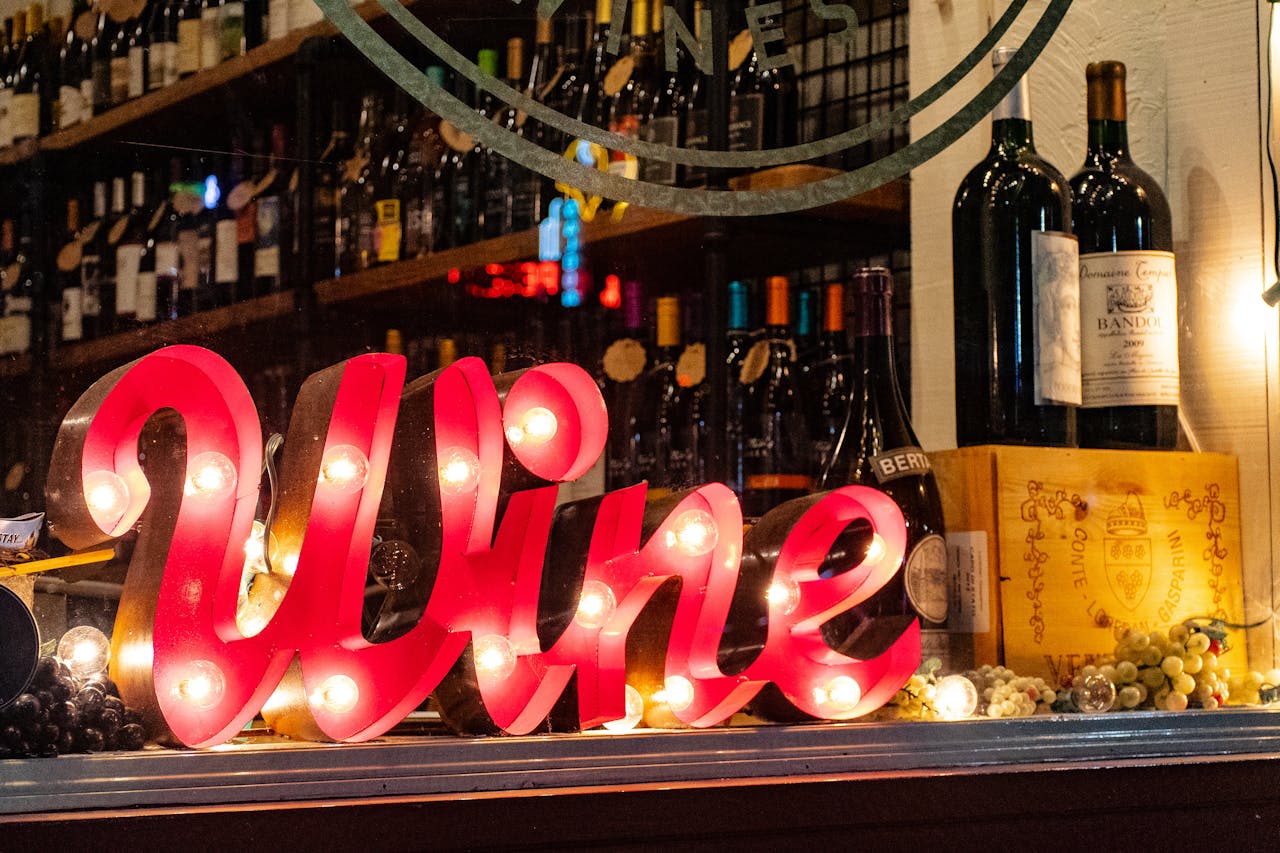 Vinbutik udstillingsvindue med en oplyst rød 'Wine' skilt og flasker vin samt vinæsker i baggrunden. Stemningsfuld og indbydende atmosfære.