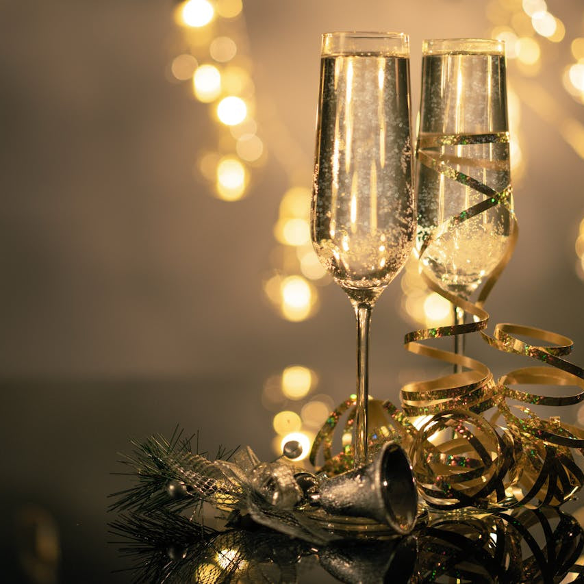 To champagneglas fyldt med bobler, dekoreret med gyldent bånd, stående på et blankt bord med julepynt og lys i baggrunden. Perfekt til festlige fejring