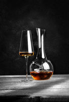 Zieher karaffel Star Mini, fyldt med en ravfarvet væske, står på et træbord foran en mørk baggrund. Ved siden af karaflen står et Zieher vinglas med samme væske