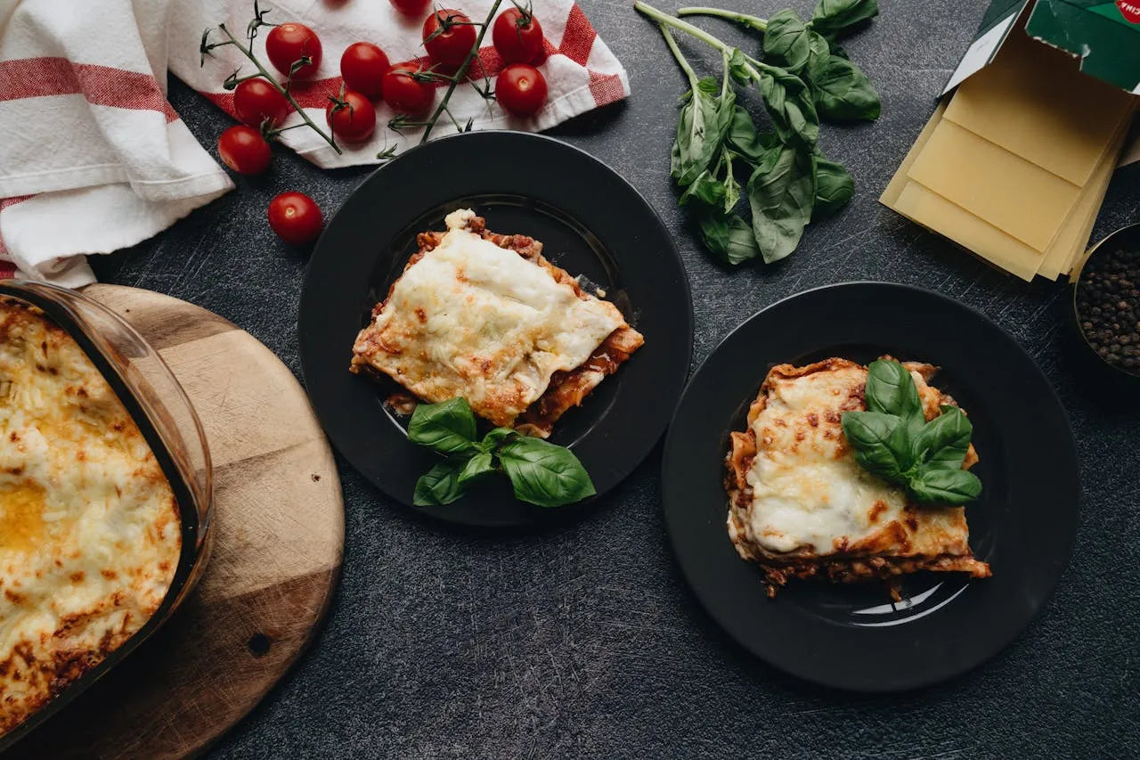 To portioner lasagne på sorte tallerkener med frisk basilikum og cherrytomater som dekoration på et køkkenbord.