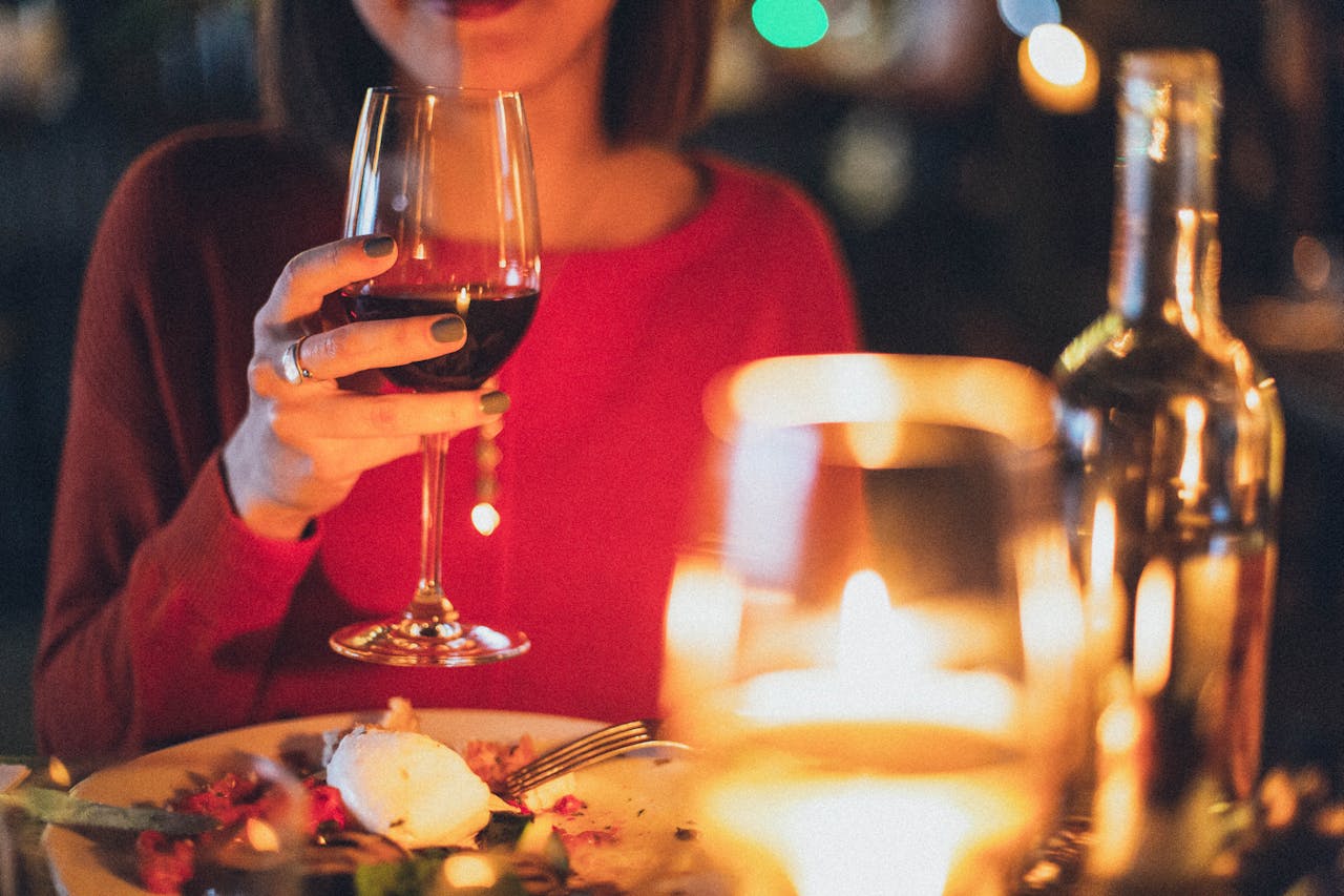 Kvinde i rød trøje nyder et glas rødvin ved et stearinlysbelyset middagsselskab. Bordet er dækket med en tallerken mad og en flaske vin