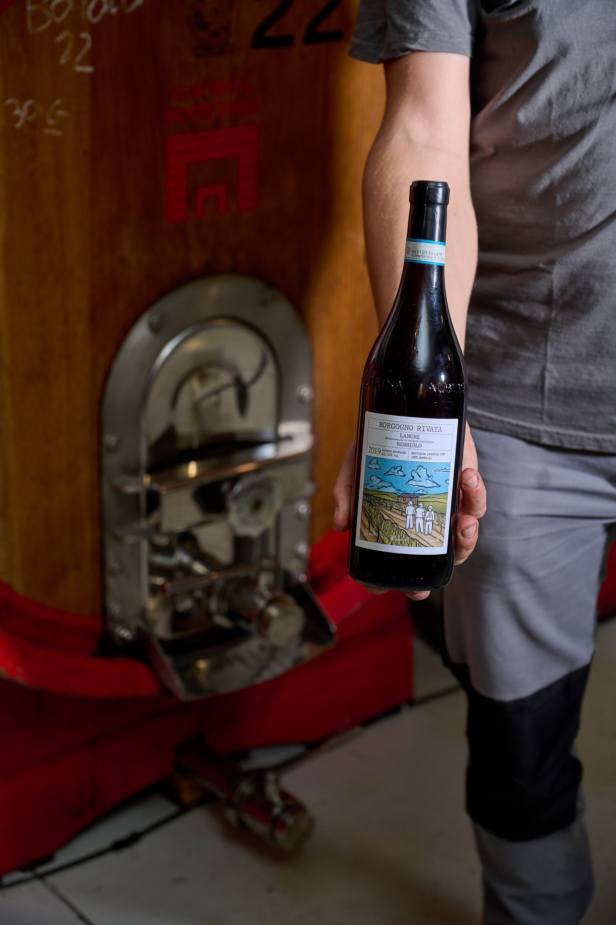 Person iført grå tøj holder en vinflaske foran en stor vintønde i en vinkælder. Fokus på flasken og etiketten.