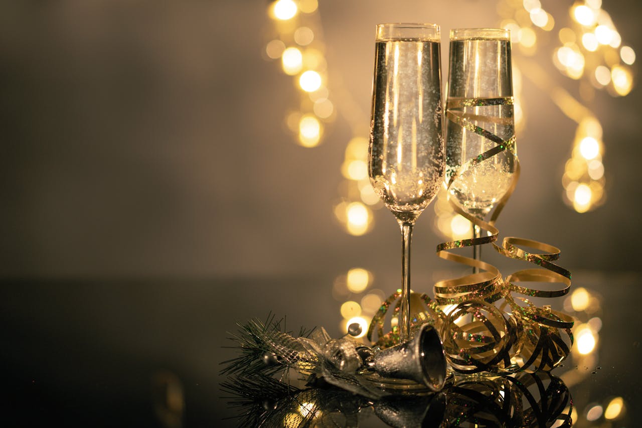 To champagneglas fyldt med bobler, dekoreret med gyldent bånd, stående på et blankt bord med julepynt og lys i baggrunden. Perfekt til festlige fejring