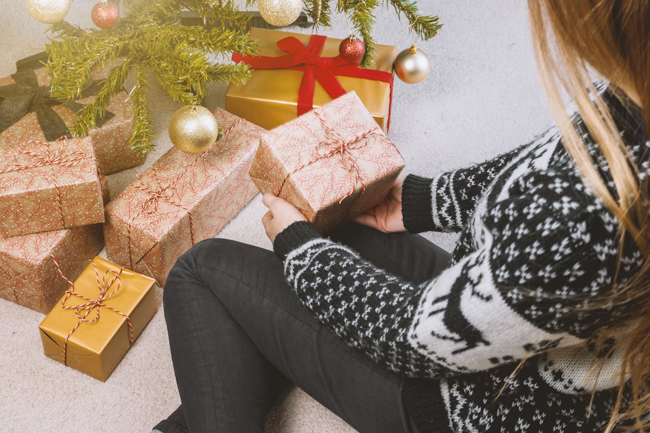 Person iført en sort og hvid julesweater sidder ved et juletræ og holder en gave, omgivet af flere festligt indpakkede julegaver